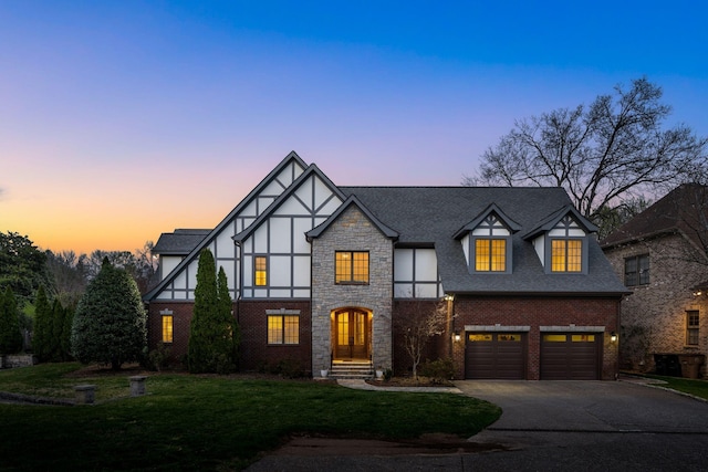 tudor home featuring roof with shingles, brick siding, a garage, a yard, and driveway