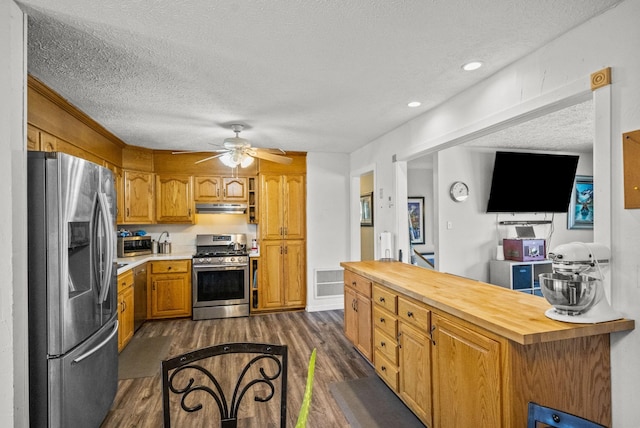 kitchen with under cabinet range hood, a textured ceiling, butcher block counters, dark wood finished floors, and appliances with stainless steel finishes