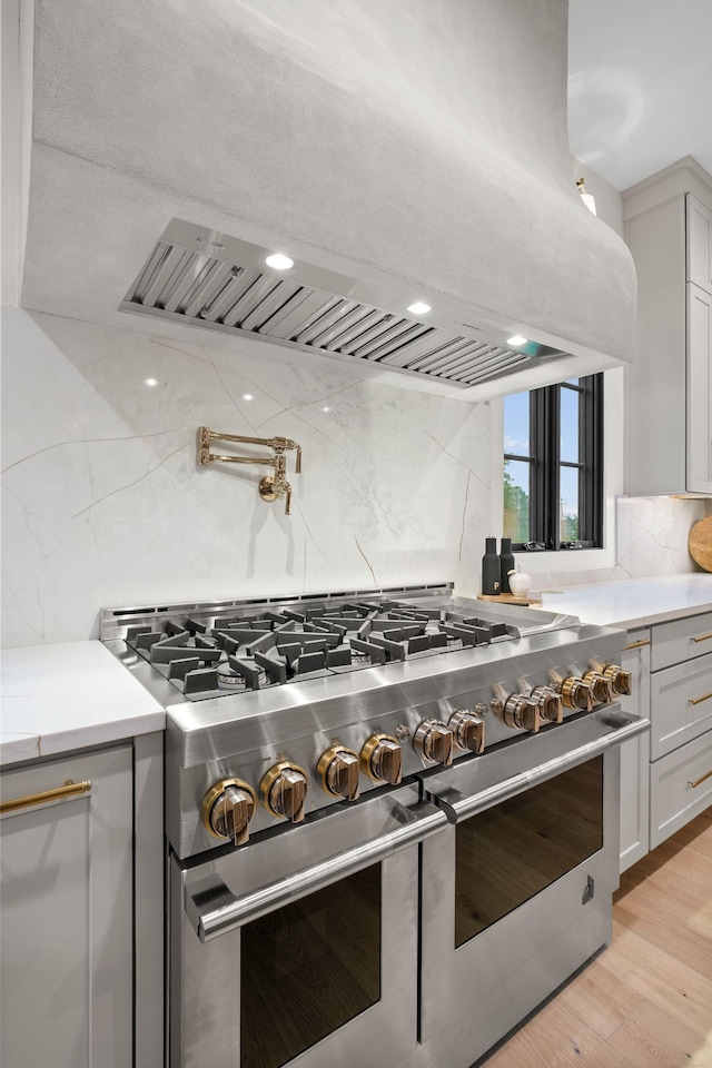 kitchen with light wood finished floors, range with two ovens, range hood, backsplash, and light countertops