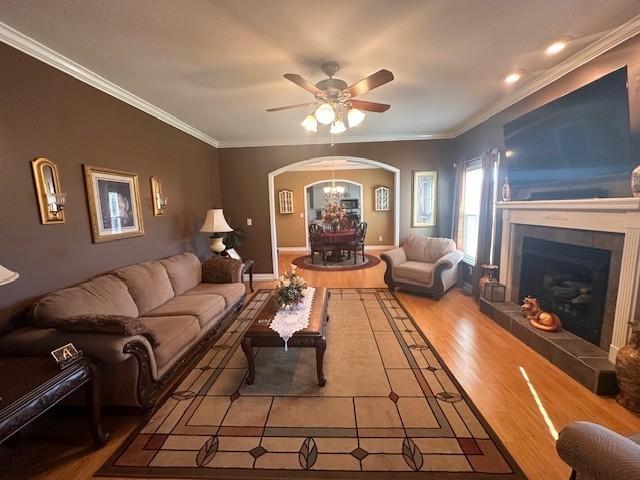 living room with ornamental molding, ceiling fan with notable chandelier, wood finished floors, arched walkways, and a fireplace