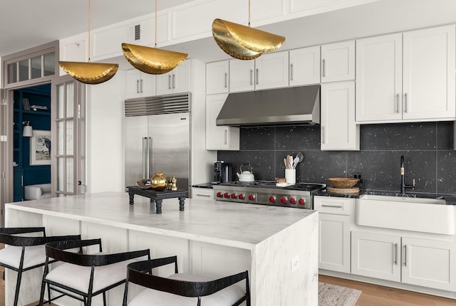 kitchen featuring under cabinet range hood, stainless steel built in fridge, a sink, white cabinetry, and backsplash