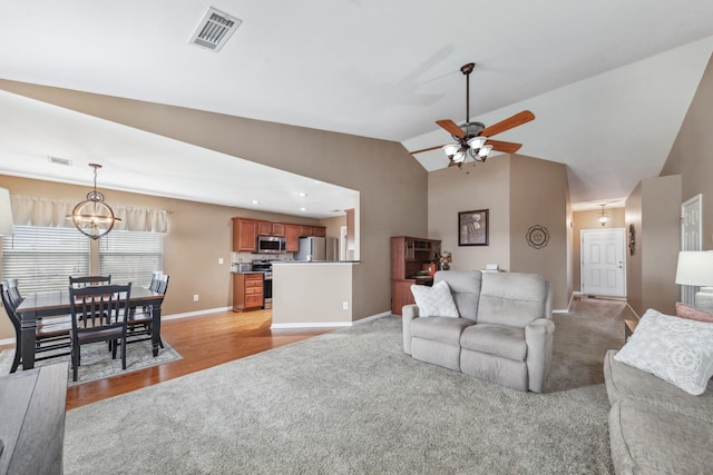 living area featuring ceiling fan with notable chandelier, visible vents, baseboards, light colored carpet, and vaulted ceiling