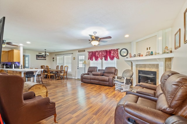 living room with ceiling fan with notable chandelier, a tiled fireplace, baseboards, light wood-style floors, and recessed lighting