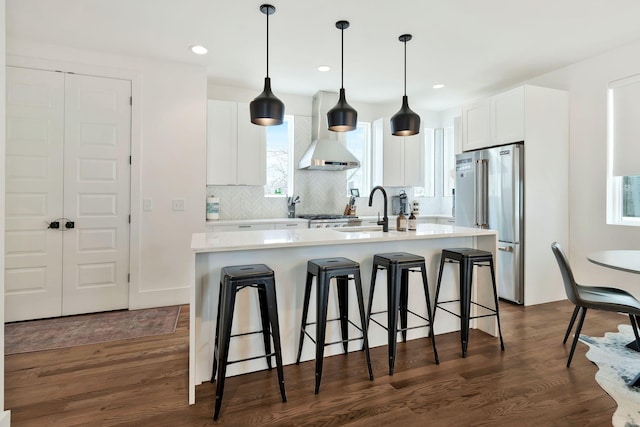 kitchen with high end fridge, a breakfast bar area, a sink, wall chimney exhaust hood, and a center island with sink