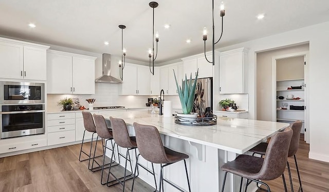 kitchen featuring wall chimney range hood, light wood-type flooring, a large island with sink, white cabinets, and stainless steel appliances