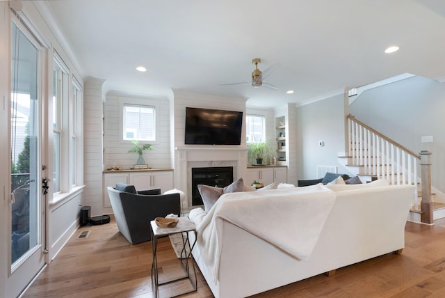 living room featuring light wood-style flooring, ceiling fan, a high end fireplace, crown molding, and stairway