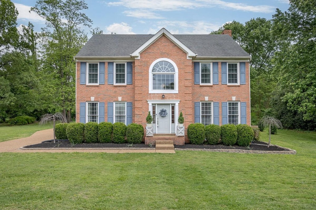 colonial-style house with a front lawn, a shingled roof, brick siding, and a chimney