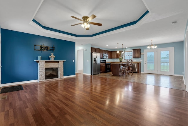 living room featuring ceiling fan with notable chandelier, dark wood-style floors, a tray ceiling, baseboards, and a stone fireplace