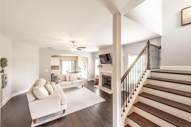 living room with a ceiling fan, baseboards, a fireplace with flush hearth, stairway, and dark wood-style floors