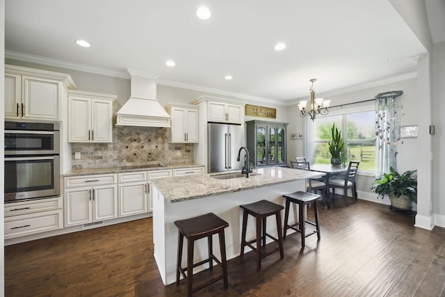 kitchen featuring an inviting chandelier, a breakfast bar area, an island with sink, appliances with stainless steel finishes, and custom range hood