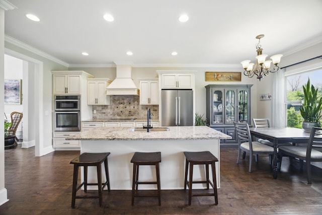 kitchen featuring a sink, stainless steel appliances, a notable chandelier, a kitchen island with sink, and custom range hood