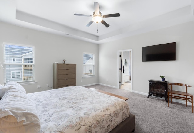 bedroom with a ceiling fan, a tray ceiling, carpet, baseboards, and a wood stove