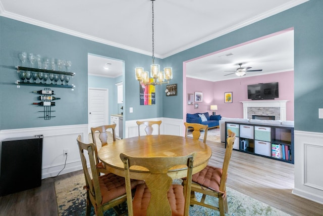 dining area with ornamental molding, wainscoting, ceiling fan with notable chandelier, wood finished floors, and a fireplace