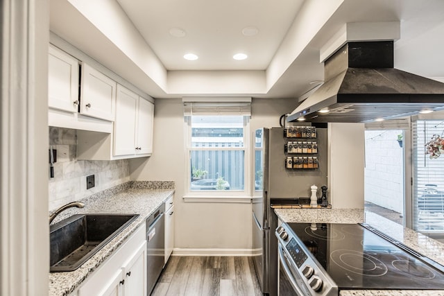 kitchen with wall chimney range hood, appliances with stainless steel finishes, wood finished floors, white cabinets, and a sink