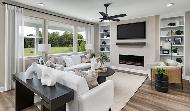 living room with baseboards, a glass covered fireplace, ceiling fan, built in features, and wood finished floors