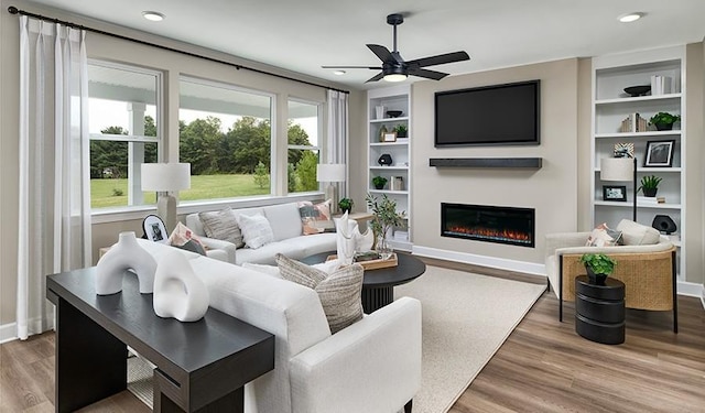 living area featuring wood finished floors, built in features, ceiling fan, baseboards, and a glass covered fireplace