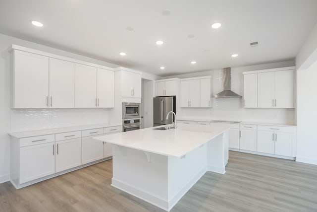 kitchen featuring a sink, a kitchen island with sink, wall chimney range hood, appliances with stainless steel finishes, and tasteful backsplash