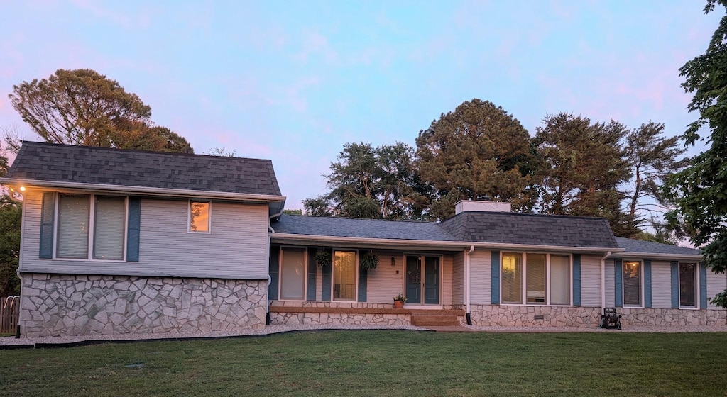 back of property at dusk with a yard, stone siding, a porch, and roof with shingles
