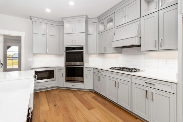 kitchen with gray cabinets, custom exhaust hood, light wood-style flooring, light countertops, and appliances with stainless steel finishes