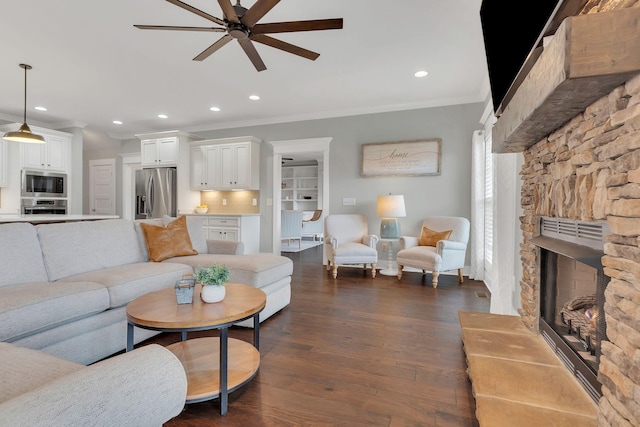 living room with a ceiling fan, recessed lighting, a stone fireplace, ornamental molding, and dark wood-type flooring