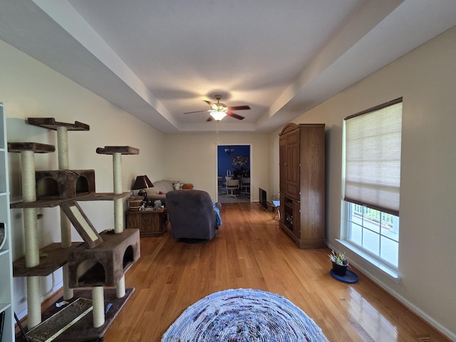 living area with a tray ceiling, light wood-type flooring, baseboards, and ceiling fan