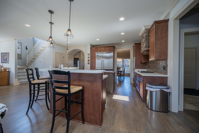 kitchen with built in refrigerator, a kitchen island with sink, dark wood-style flooring, light countertops, and ornamental molding