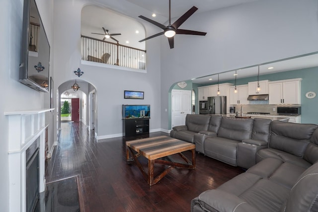 living area featuring arched walkways, a ceiling fan, dark wood-type flooring, a towering ceiling, and baseboards