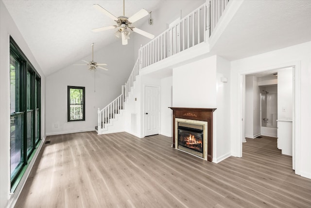unfurnished living room with ceiling fan, light wood-style flooring, high vaulted ceiling, a glass covered fireplace, and stairway