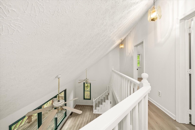 hall featuring a textured ceiling, light wood-style floors, vaulted ceiling, baseboards, and an upstairs landing