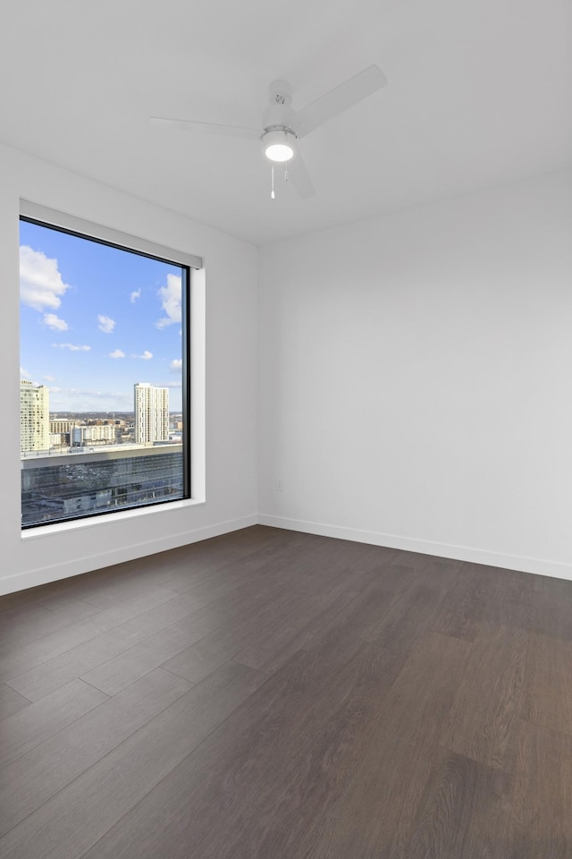 unfurnished room with dark wood-style floors, a ceiling fan, baseboards, and a view of city