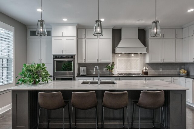 kitchen with a sink, premium range hood, stainless steel appliances, backsplash, and a center island with sink