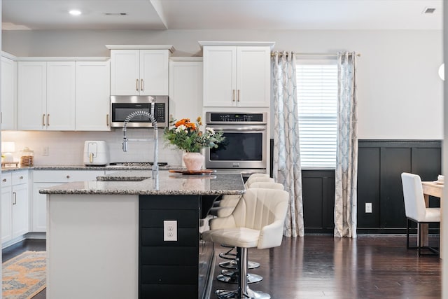 kitchen with appliances with stainless steel finishes, white cabinets, light stone countertops, a kitchen bar, and wainscoting