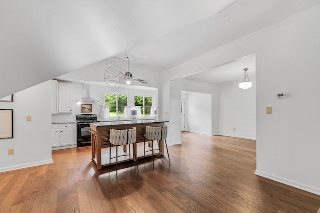 kitchen with black range with electric stovetop, wall chimney range hood, a kitchen breakfast bar, white cabinets, and wood finished floors