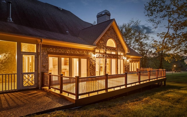 back of house with a wooden deck, stone siding, a chimney, a lawn, and roof with shingles