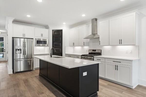 kitchen featuring stainless steel appliances, wall chimney exhaust hood, a sink, light wood-style floors, and an island with sink