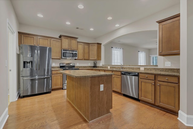 kitchen featuring stainless steel appliances, arched walkways, light wood-style floors, brown cabinetry, and a center island