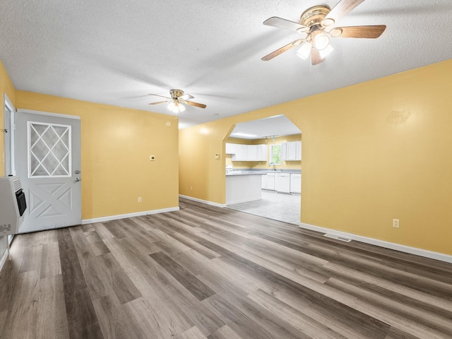 unfurnished living room with ceiling fan, arched walkways, light wood-style flooring, a textured ceiling, and baseboards