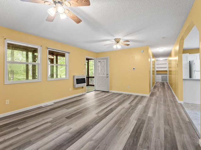 unfurnished living room featuring ceiling fan, heating unit, a textured ceiling, wood finished floors, and baseboards