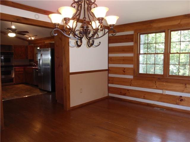unfurnished dining area with a chandelier, dark wood finished floors, wood walls, baseboards, and a ceiling fan