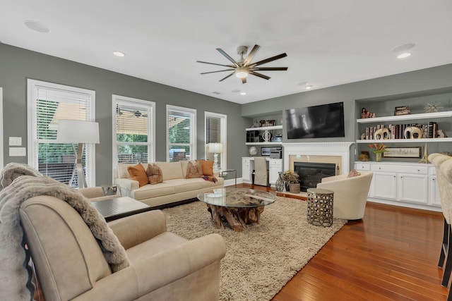 living area featuring ceiling fan, a glass covered fireplace, dark wood-style floors, built in shelves, and recessed lighting