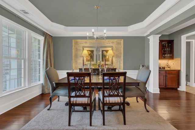dining space with a raised ceiling, a chandelier, crown molding, dark wood-type flooring, and ornate columns