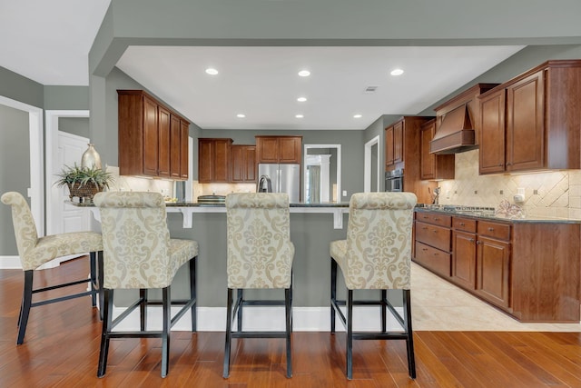 kitchen with premium range hood, stainless steel appliances, a kitchen bar, backsplash, and light wood finished floors