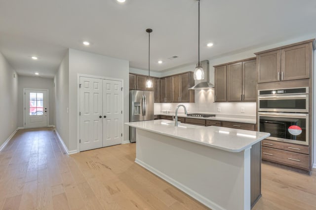 kitchen with appliances with stainless steel finishes, wall chimney exhaust hood, light wood finished floors, a kitchen island with sink, and a sink