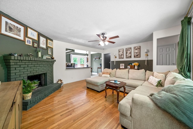 living room featuring light wood-style floors, ceiling fan, a textured ceiling, a brick fireplace, and baseboards