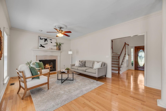 living room with a tile fireplace, plenty of natural light, stairs, a ceiling fan, and crown molding