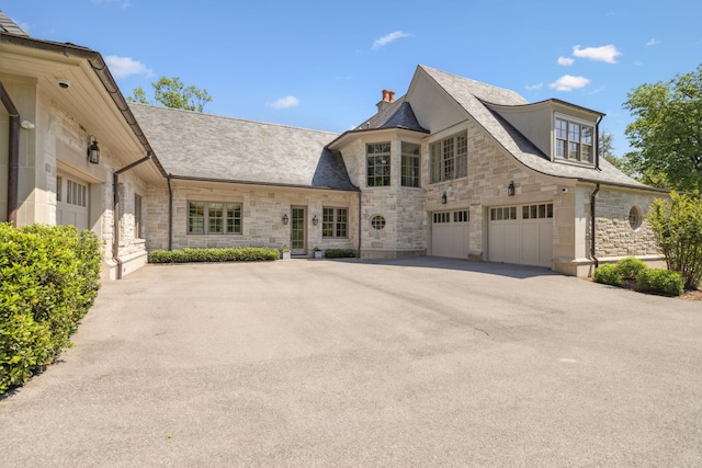 french country home featuring stone siding, a garage, driveway, and a chimney