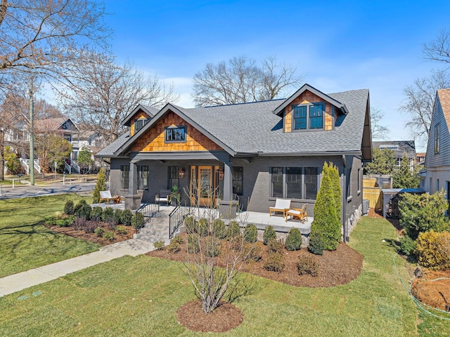 craftsman-style home featuring roof with shingles, covered porch, a front lawn, and brick siding