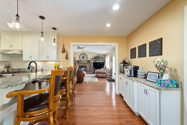 kitchen featuring under cabinet range hood, ceiling fan, a glass covered fireplace, light wood-style floors, and a kitchen breakfast bar