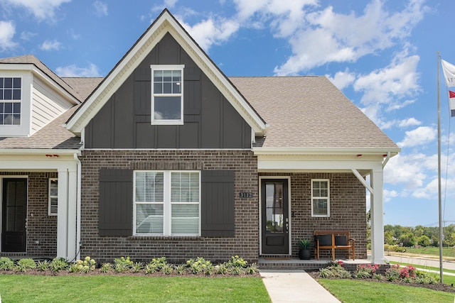 view of front of property featuring brick siding, board and batten siding, covered porch, roof with shingles, and a front yard