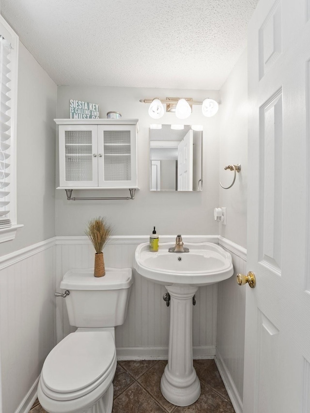 half bathroom with a textured ceiling, toilet, wainscoting, tile patterned flooring, and a sink